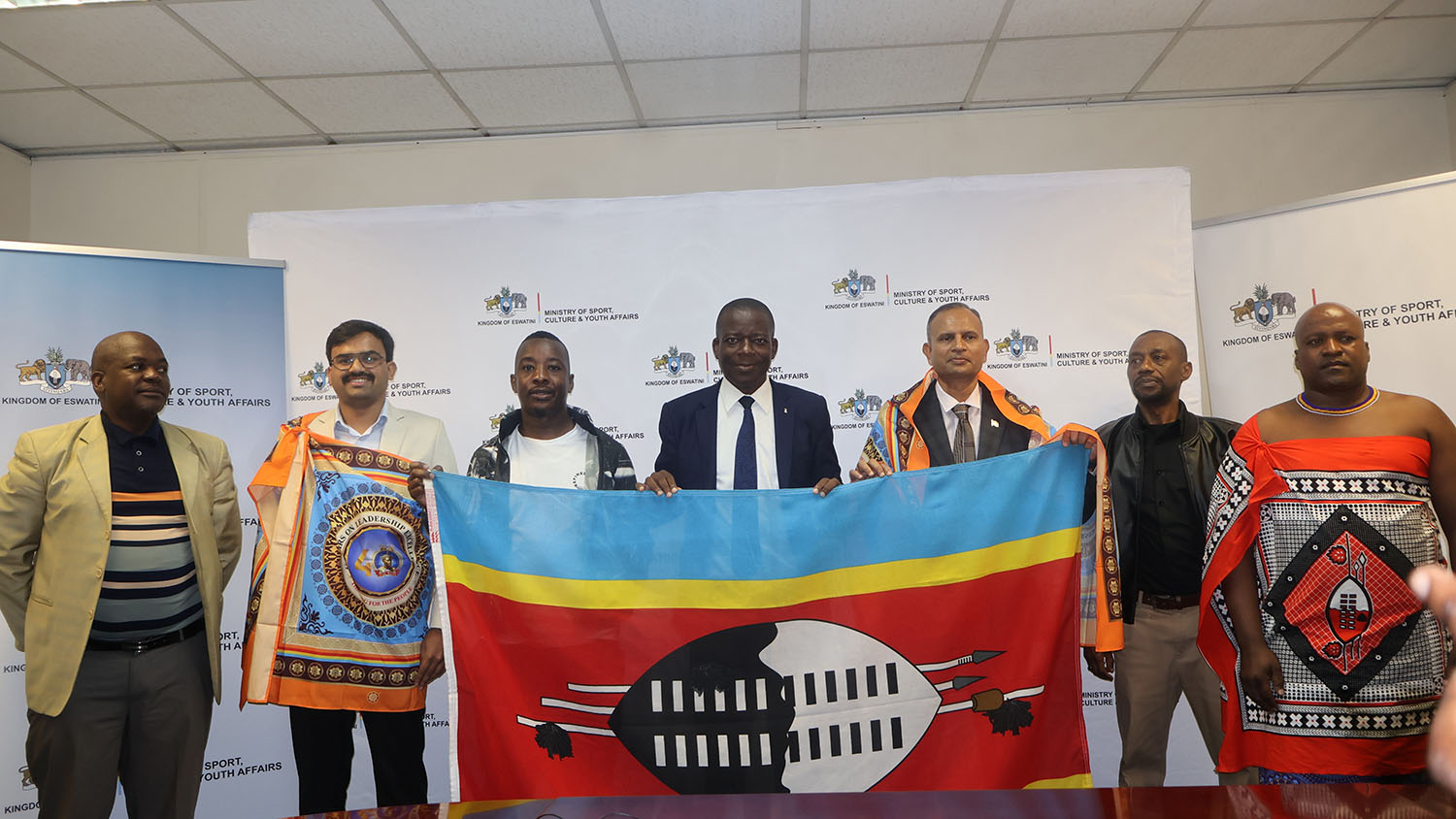 Ministry of Sports, Culture and Youth Affairs, Bongani Ndzima (C), officially handing the Eswatini flag to Honest Mhlanga during his farewell ahead of his cultural exchange trip to India, joined by the Indian High Commissioner M. Ram Prasad along with the CEO of the Ministry, Stanley Dlamini and other officials.